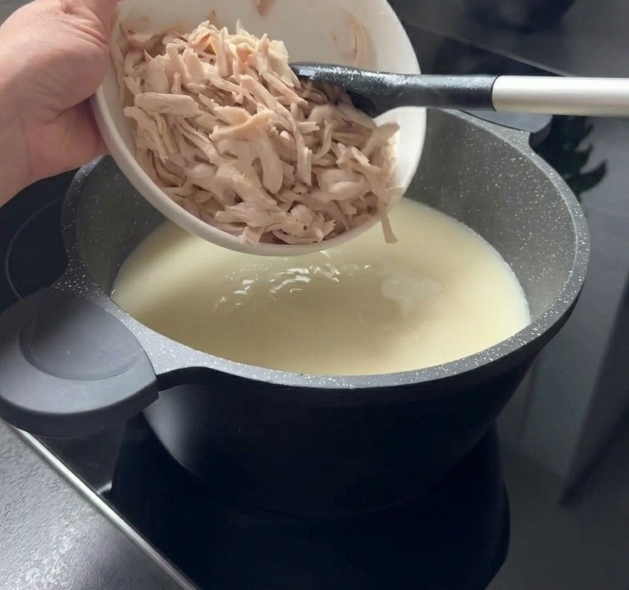 Gezupfte Stücke von gegartem Hähnchenfleisch werden aus einer weißen Schüssel in einen Topf mit cremiger weißer Suppe gegeben.