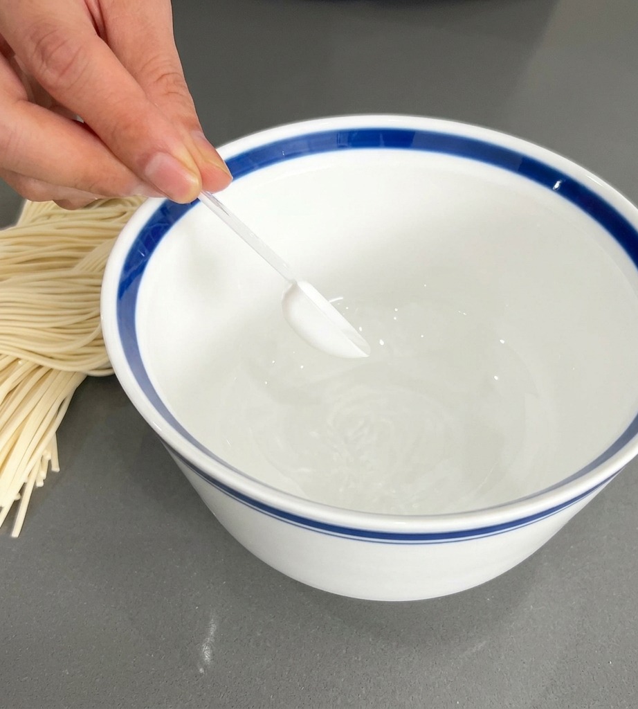 Eine Hand gibt mit einem kleinen transparenten Löffel weißes Würzpulver in eine weiße Schale mit blauem Rand.