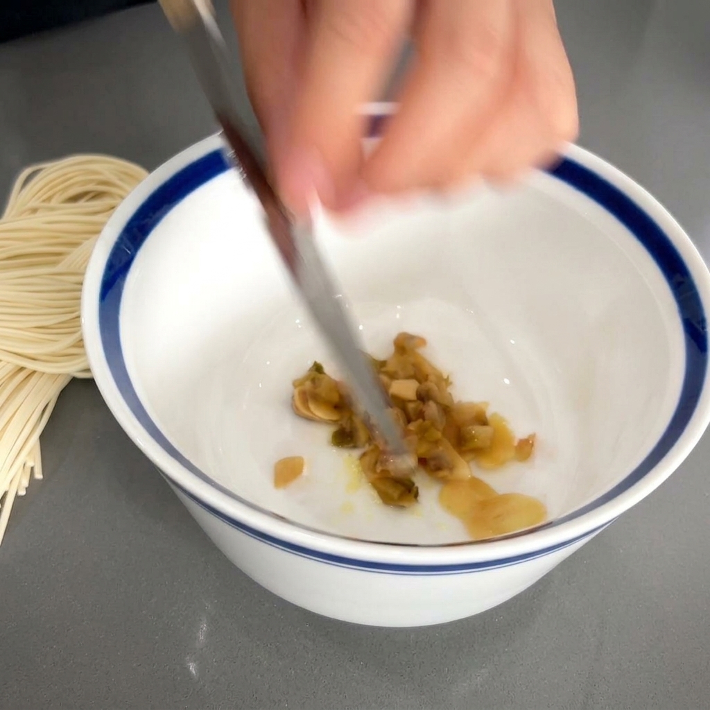 Eine Messerklinge schiebt fein gehacktes asiatisches eingelegtes Gemüse in die Schale zum Knoblauch.