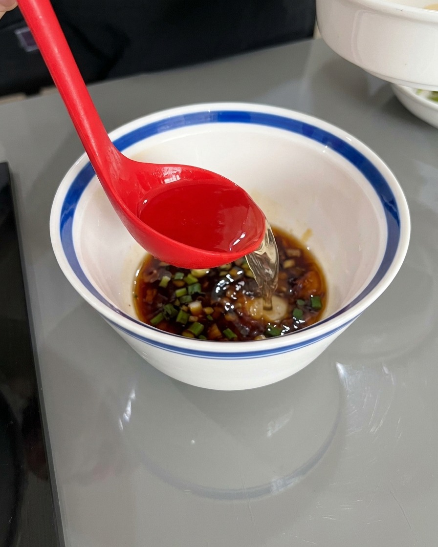 Eine rote Schöpfkelle gießt klares heißes Wasser in die weiße Schale mit Sojasauce und Gewürzen.