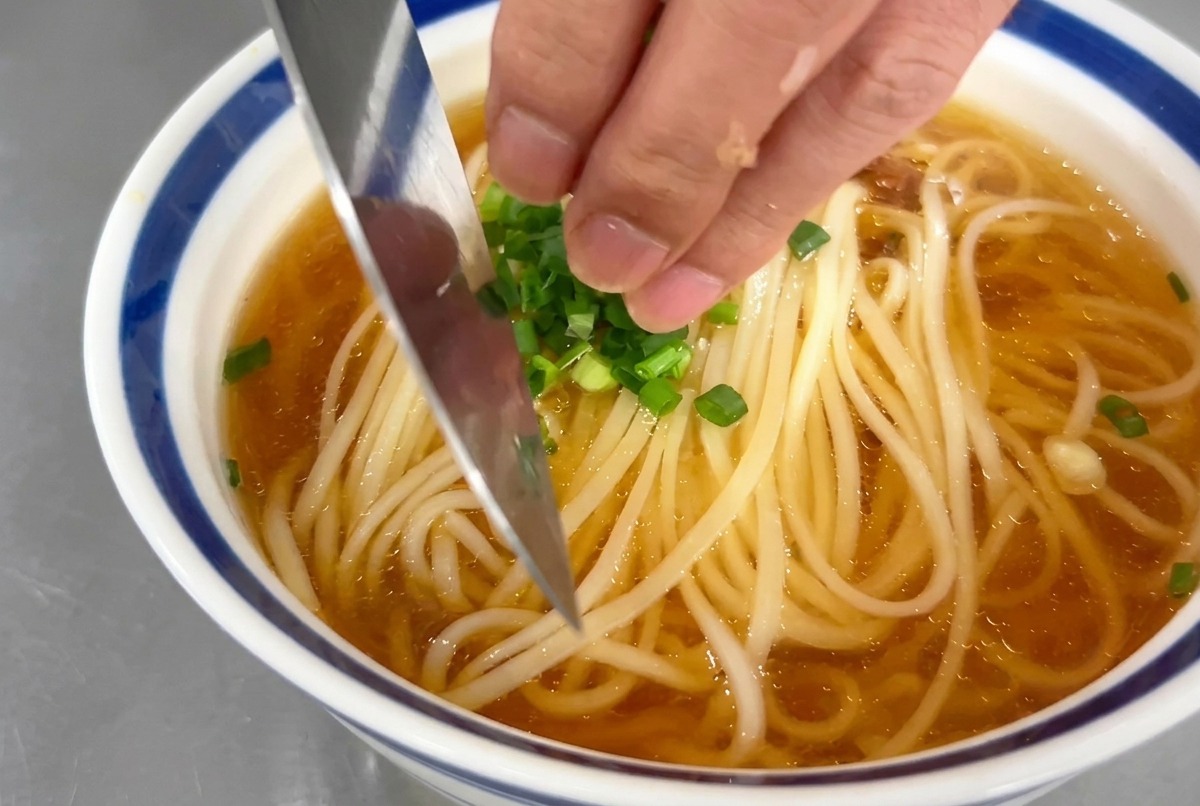 Eine Hand streut frische Frühlingszwiebelringe über eine Schale Nudelsuppe.