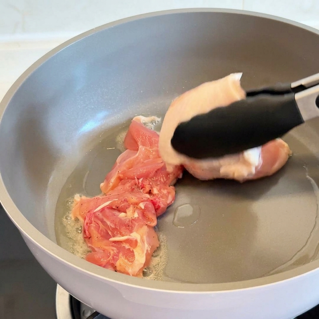 Eine Grillzange legt ein rohes Hähnchenschenkelstück mit der Hautseite nach unten in eine graue, beschichtete Pfanne mit heißem Öl.