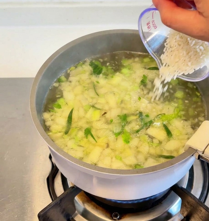 Weißer Reis wird aus einem transparenten Messbecher in einen Topf mit köchelnder Suppe und sichtbarem Gemüse geschüttet.