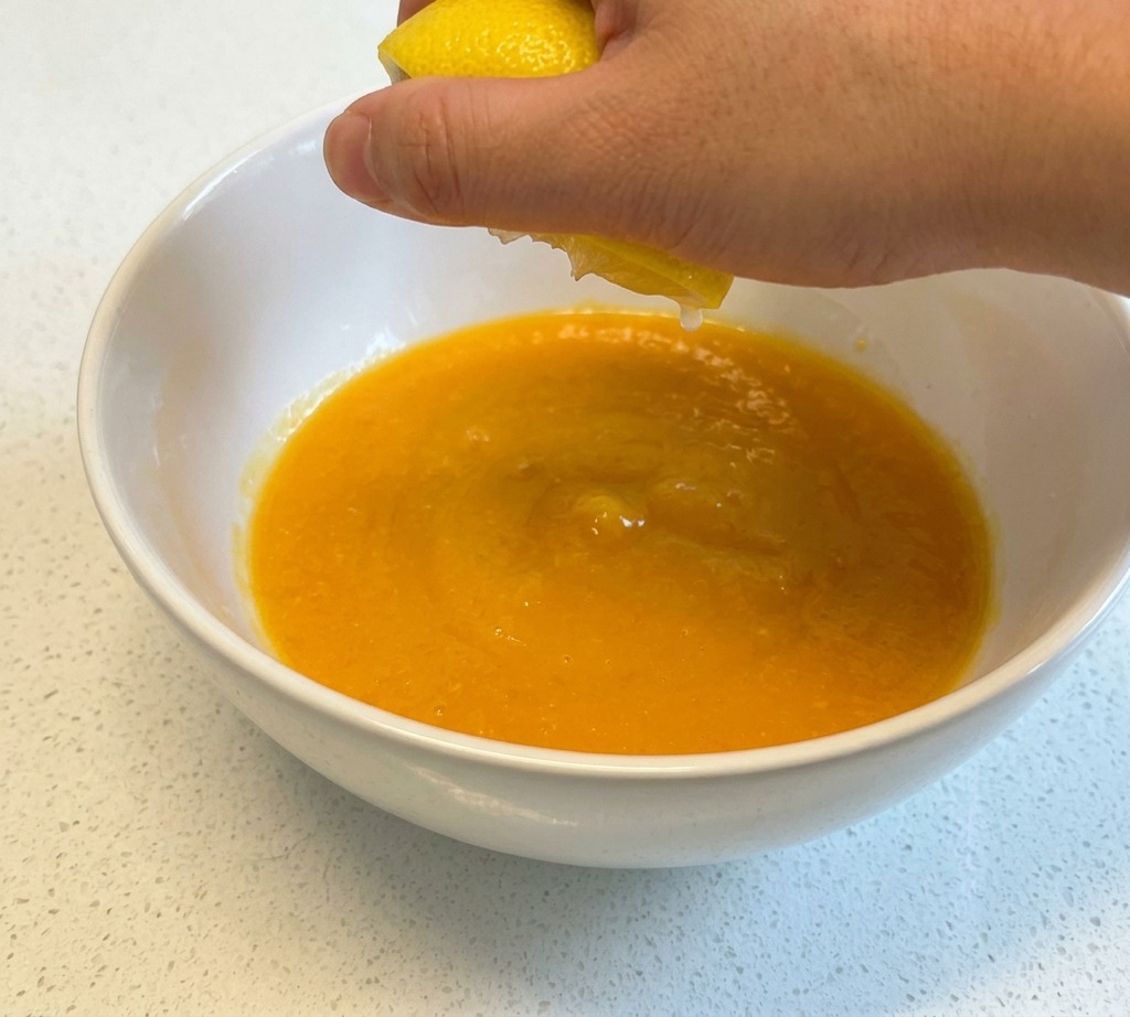 Eine Hand presst den Saft einer gelben Zitronenhälfte in eine weiße Keramikschüssel mit verquirlten Eiern.