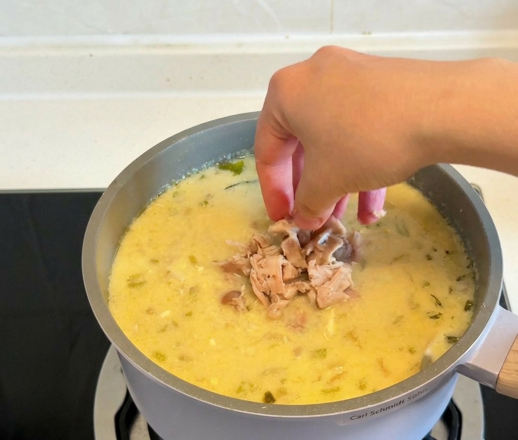 Hände geben das gezupfte, gegarte Hähnchenfleisch zurück in einen Topf mit der cremig-gelb gebundenen Suppe.