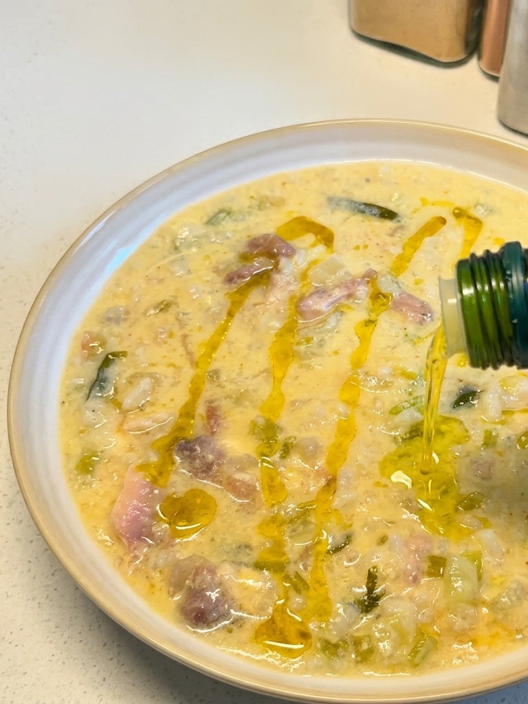 Goldenes Olivenöl wird aus einer grünen Flasche über eine angerichtete Schüssel mit cremiger Zitronen-Hühnersuppe geträufelt.