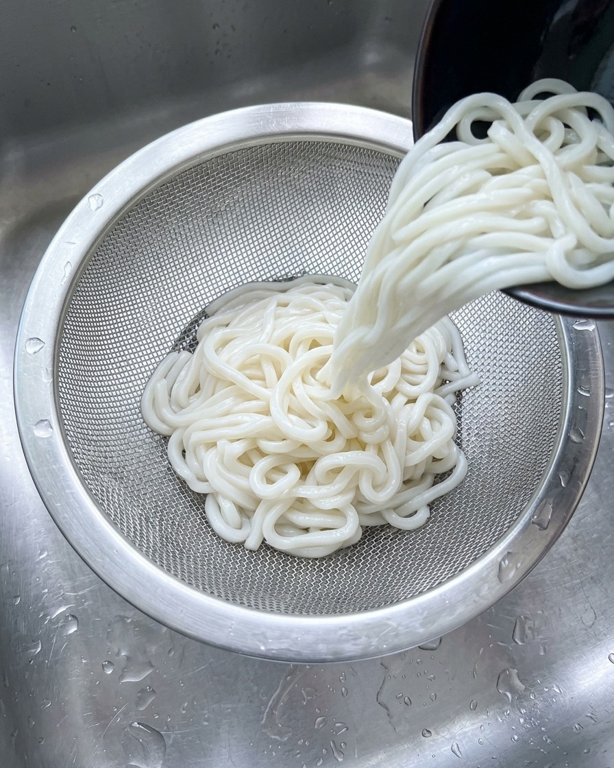 Weiße Udon-Nudeln werden in einem Sieb aus Edelstahl unter einem kalten Wasserstrahl in der Spüle abgespült.