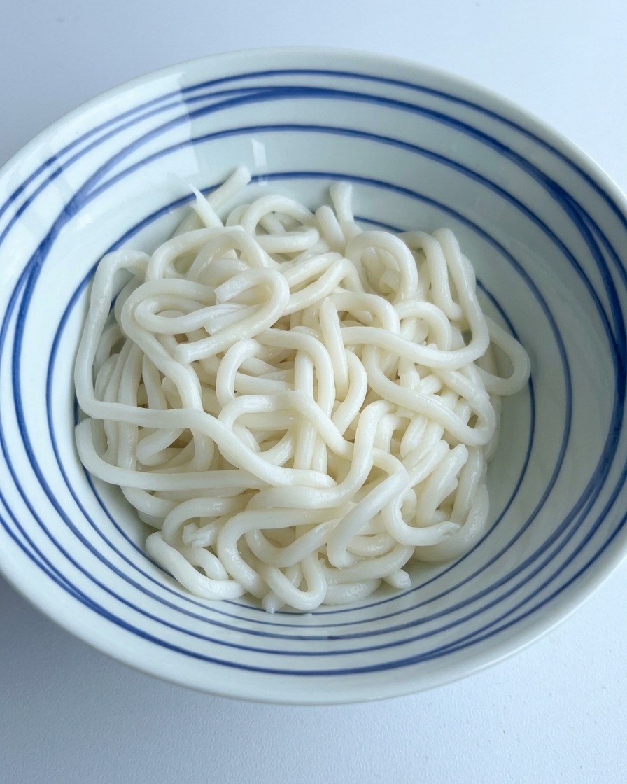 Dunkle Sojasauce und Gewürze werden über einen Haufen weißer Udon-Nudeln in einer blau gestreiften Schale gegossen.