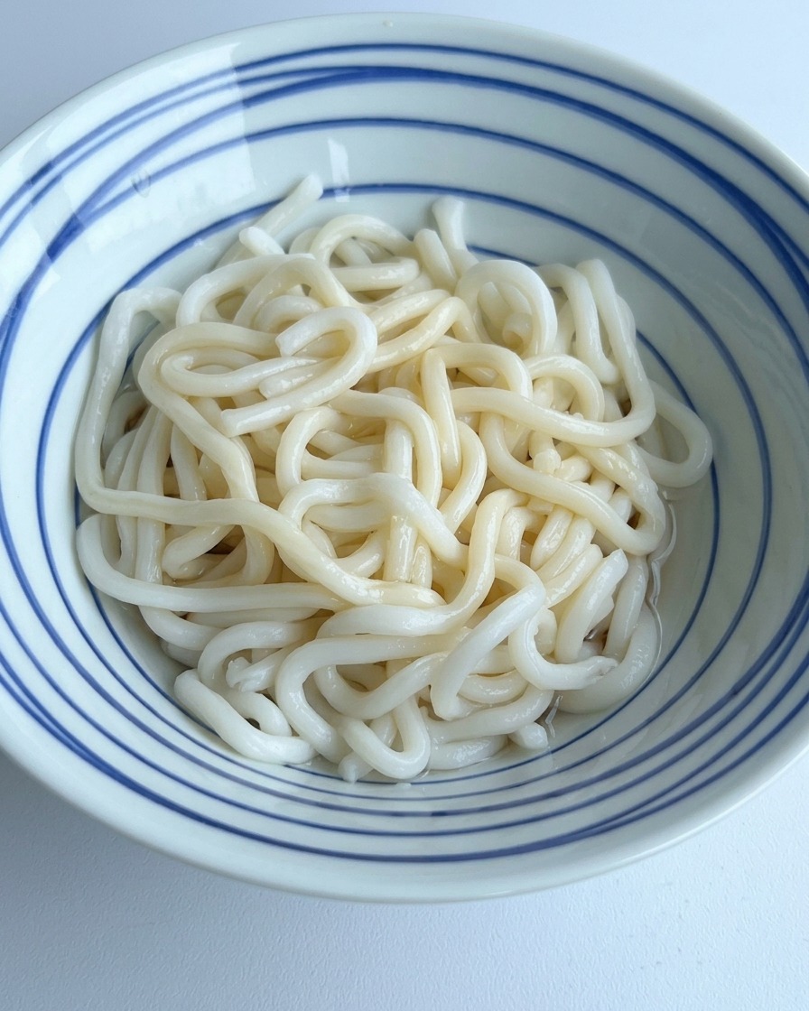 Klares Wasser wird in eine Schale mit Udon-Nudeln gegossen und vermischt sich mit den dunklen Gewürzen am Boden.