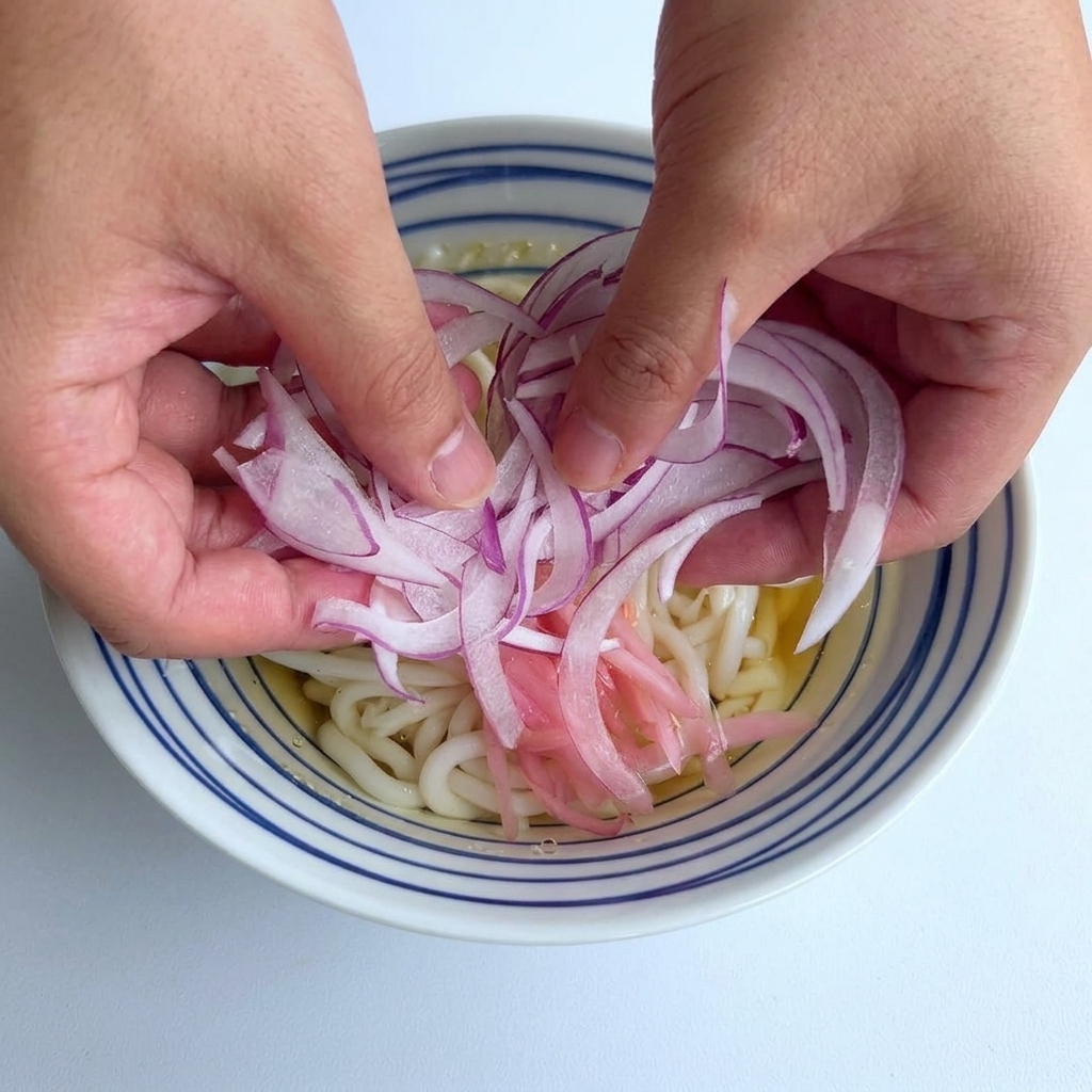 Zwei Hände streuen eine Portion dünn geschnittener purpurroter Zwiebeln in die Mitte einer Schale mit Udon-Nudeln.