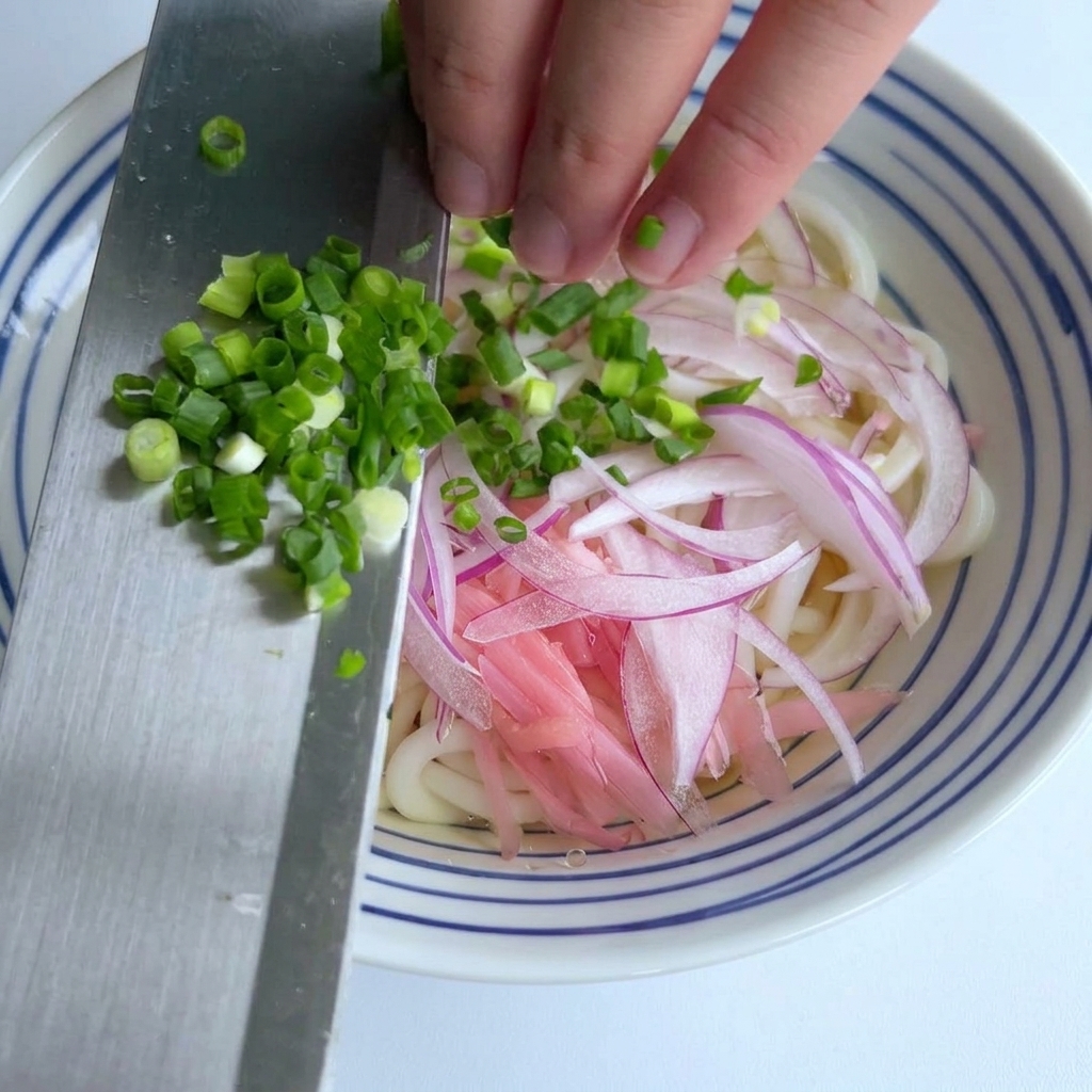 Fein gehackte Frühlingszwiebeln werden von der Seite eines Metallmessers auf eine Schale mit Udon-Nudeln und roten Zwiebeln geschoben.