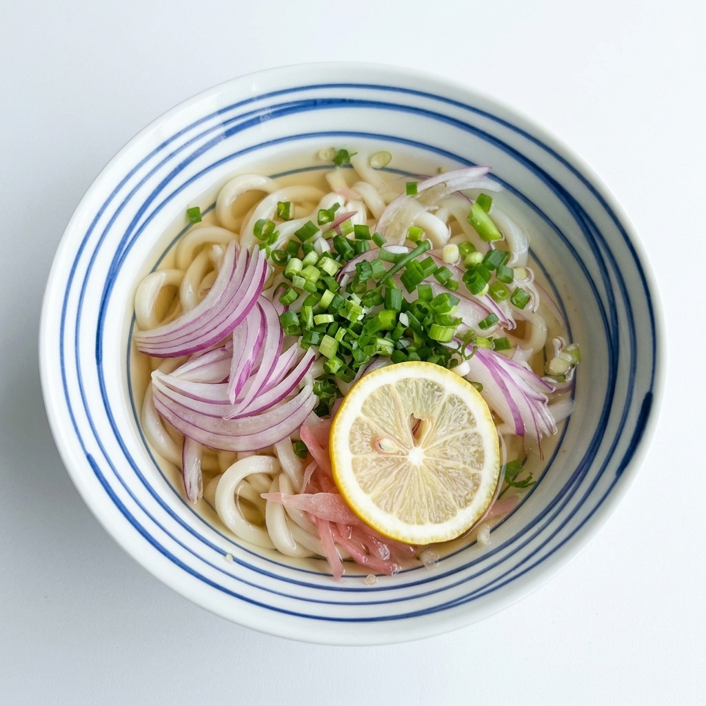 Draufsicht auf eine Schale mit Udon-Nudeln in klarer Brühe, garniert mit roten Zwiebeln und Frühlingszwiebeln, in deren Mitte eine einzelne gelbe Zitronenscheibe liegt.