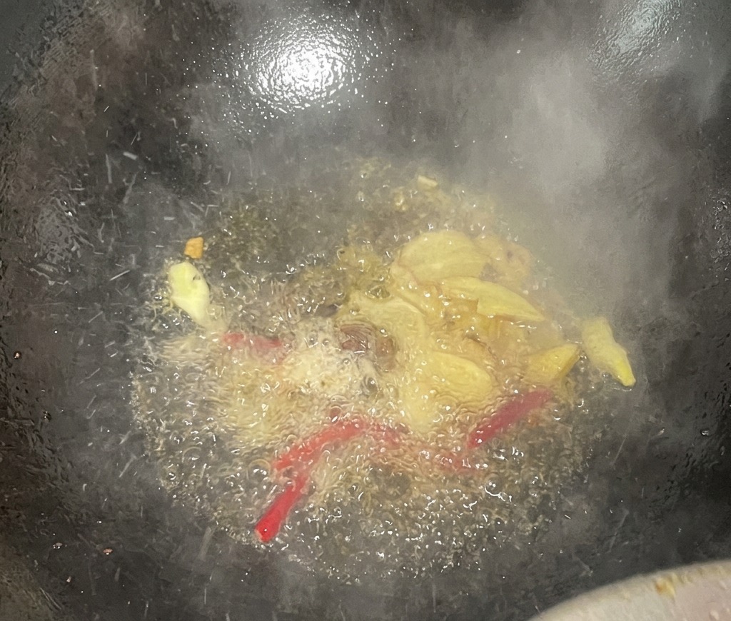 Ingwerscheiben und getrocknete rote Chilis brutzeln in heißem Öl in einem dunklen Wok und setzen aromatischen Dampf frei.