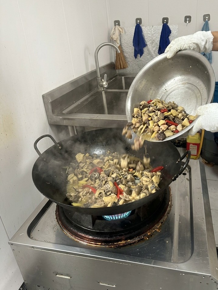Eine person gießt klares, kochendes Wasser aus einem Gefäß in einen dampfenden Wok mit angebratenem Huhn und Gewürzen.