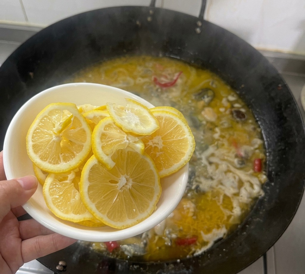 Eine Hand hält eine weiße Schüssel mit dünnen Zitronenscheiben über einen kochenden Wok mit goldener Hühnerbrühe.