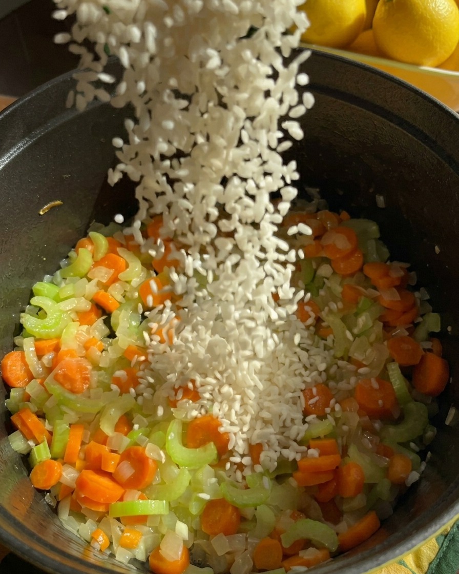 Ein Strahl weißer Jasminreis wird in einen dunklen Topf mit angedünsteten Karotten, Sellerie und Zwiebeln gegossen.
