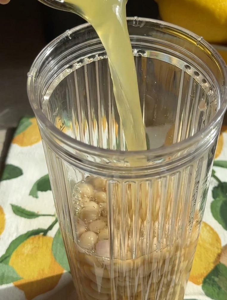 Eine gelbliche Flüssigkeit wird aus einem Glas in einen Mixerbecher gegossen, der mit ganzen weißen Bohnen gefüllt ist.