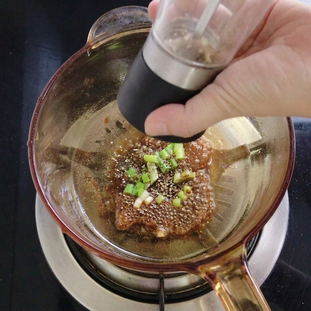 Eine Person mahlt mit einer schwarzen Gewürzmühle frischen Sichuan-Pfeffer über die Zwiebel-Öl-Mischung im Topf.
