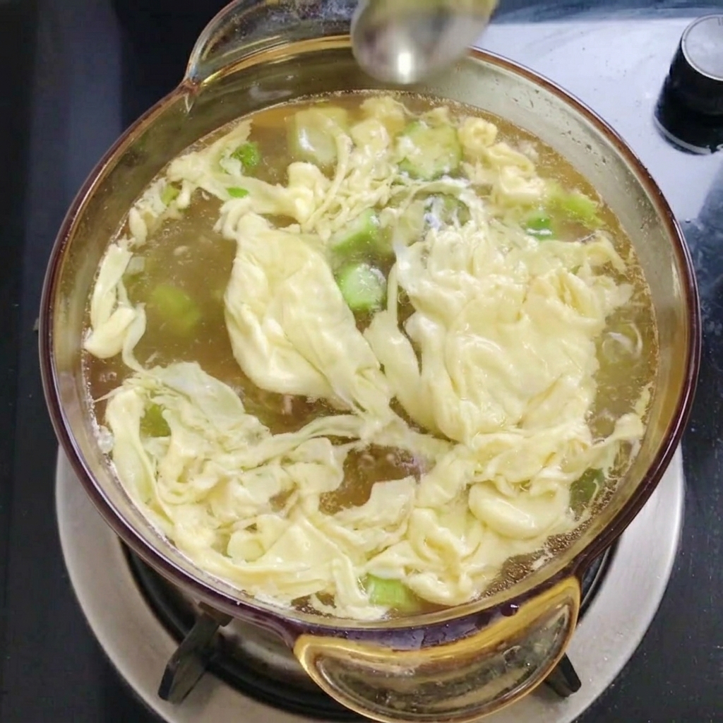 Ein Metalllöffel streut feines weißes Salz in die Suppe mit Eifäden und Okraschoten.