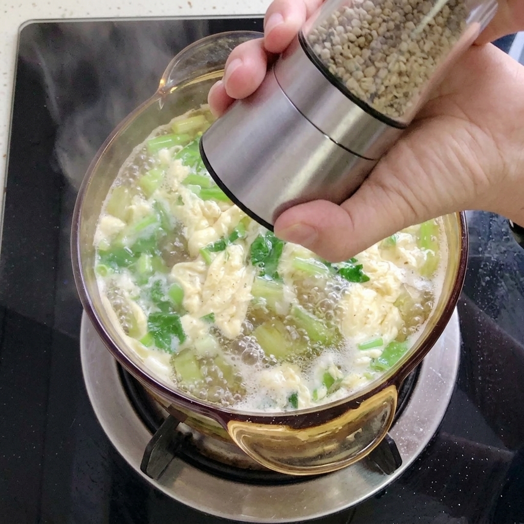 Eine Hand mahlt mit einer Edelstahl-Pfeffermühle weißen Pfeffer über die fertige Suppe voller Kräuter und Eifäden.