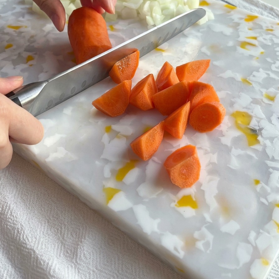 Ein Küchenmesser schneidet eine leuchtend orangefarbene Karotte auf einem weiß-gelb gemusterten Schneidebrett in Stücke, daneben liegt ein Haufen gewürfelte Zwiebeln.