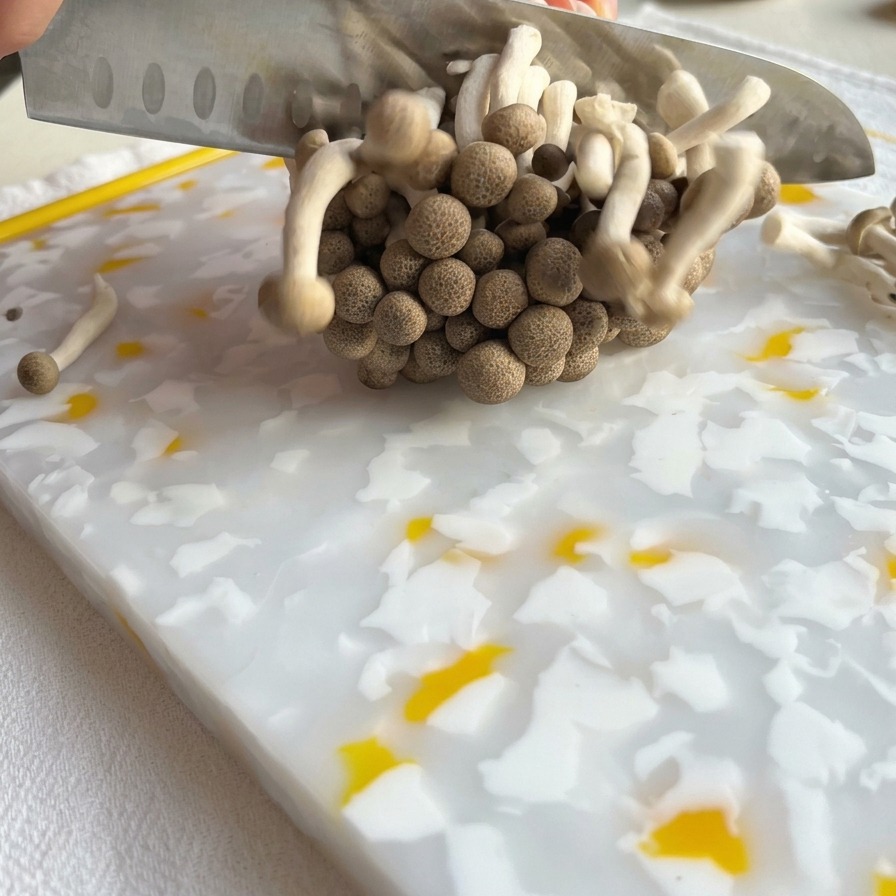 Ein Küchenmesser schneidet die Wurzelbasis eines Büschels brauner Shimeji-Pilze auf einem gemusterten Schneidebrett ab.