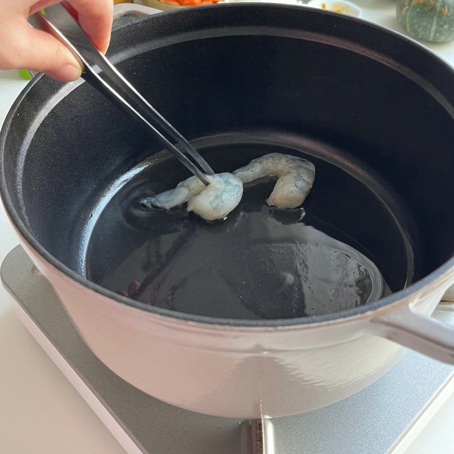 Rohe Garnelen werden mit einer Metallpinzette zum Anbraten in heißes Öl in einen schwarzen Topf gegeben.