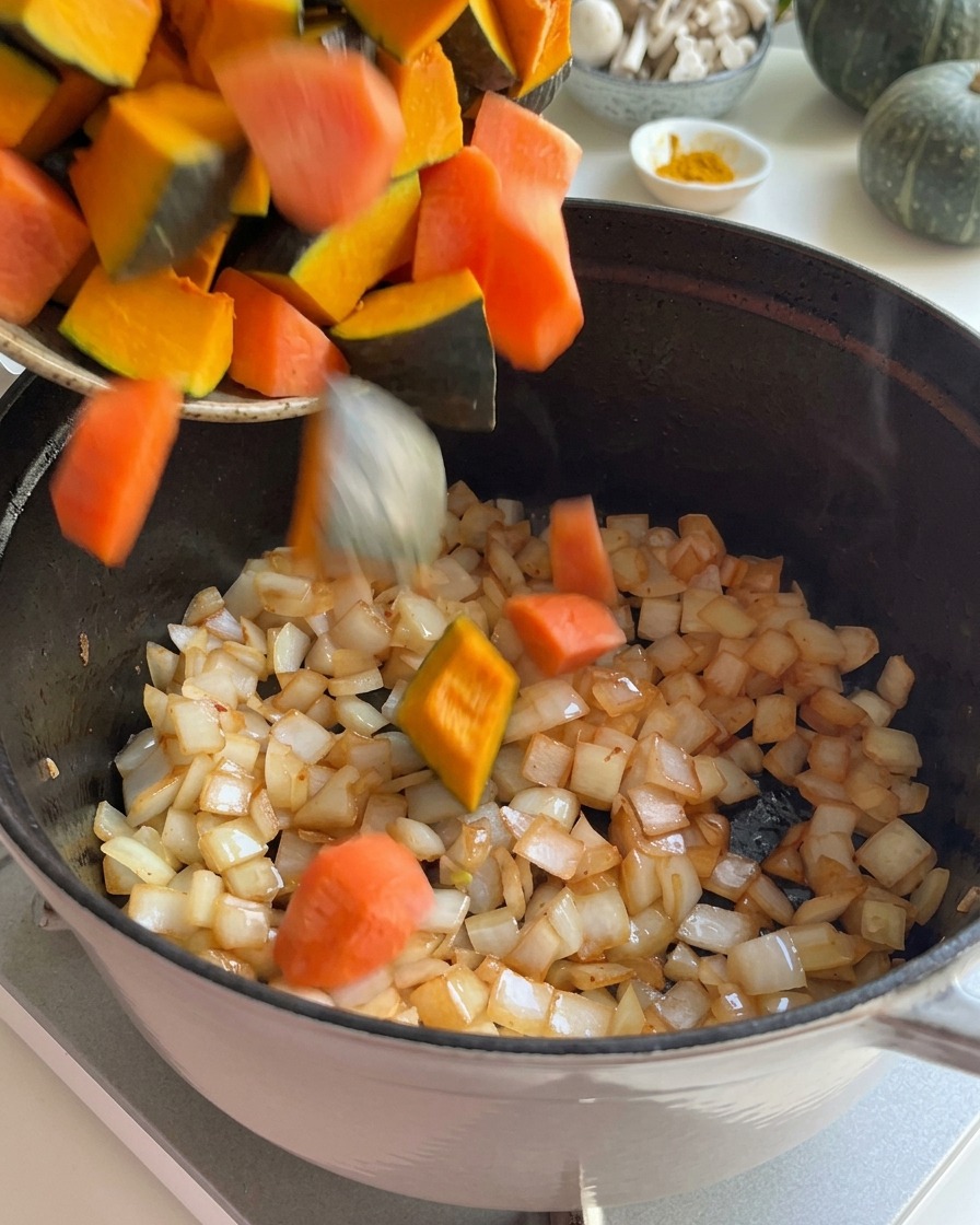 Frische Kürbisstücke und geschnittene Karotten werden zu den angeschwitzten Zwiebeln in einen Topf gegeben.