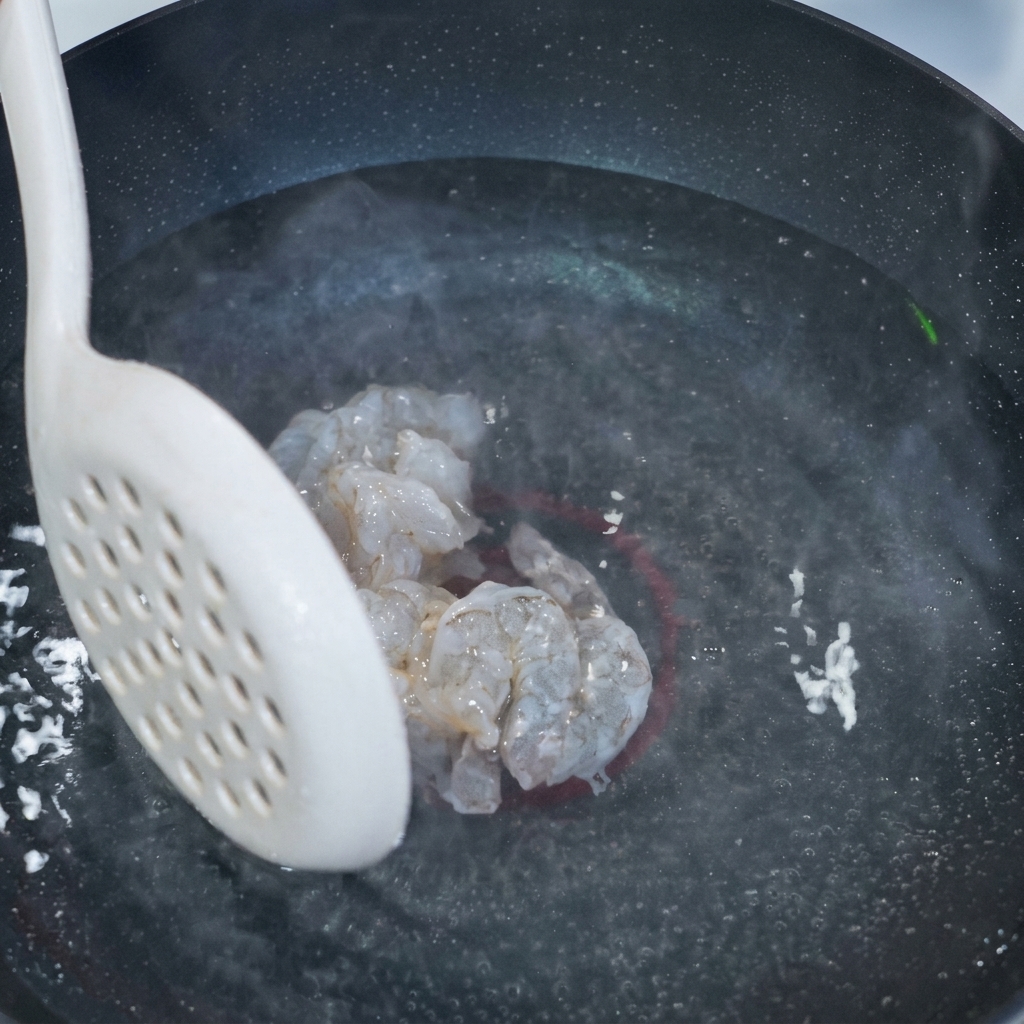 Ein weißer Schaumlöffel hält mehrere Stücke glasiges, mariniertes Garnelenfleisch über einem Topf mit dampfendem Wasser.