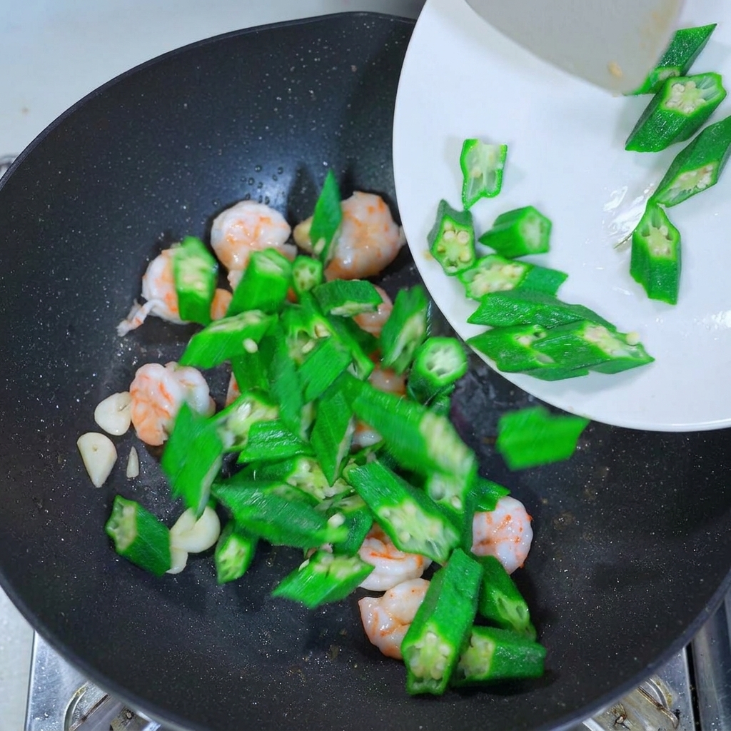 Kräftig grüne Okrascheiben werden von einem weißen Teller in einen Wok mit Garnelen und Knoblauch gegeben.