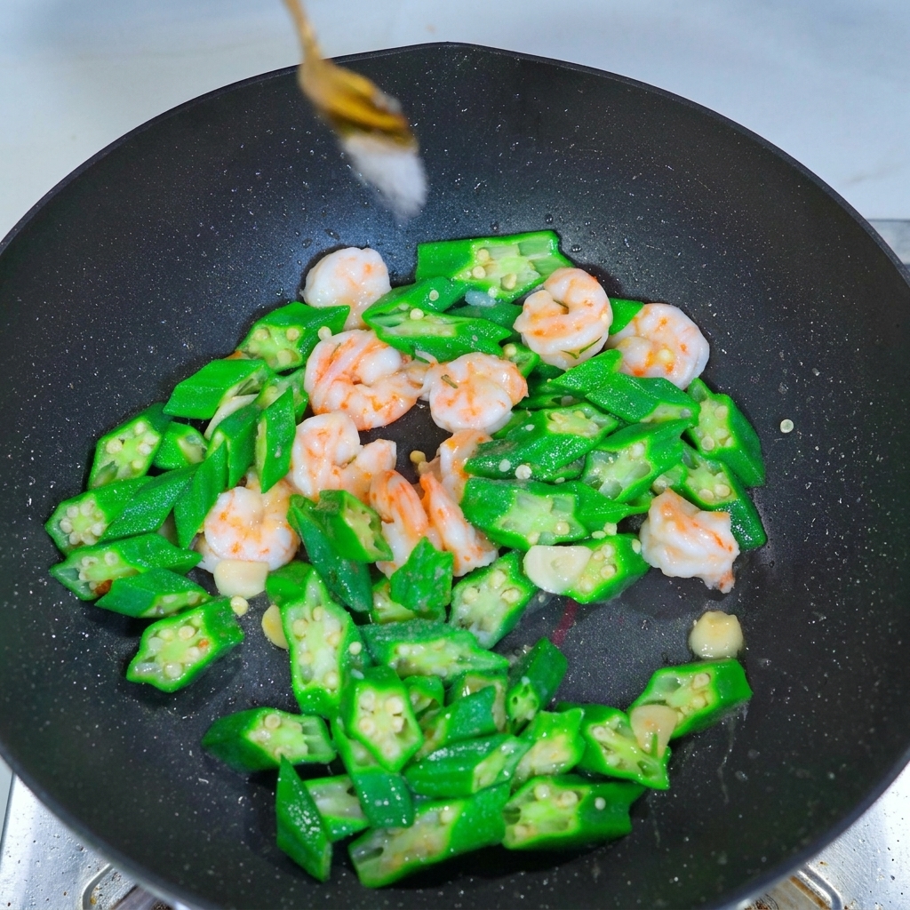 Ein Metalllöffel streut weißes Gewürz über eine Mischung aus grünen Okrascheiben und rosa Garnelen im Wok.