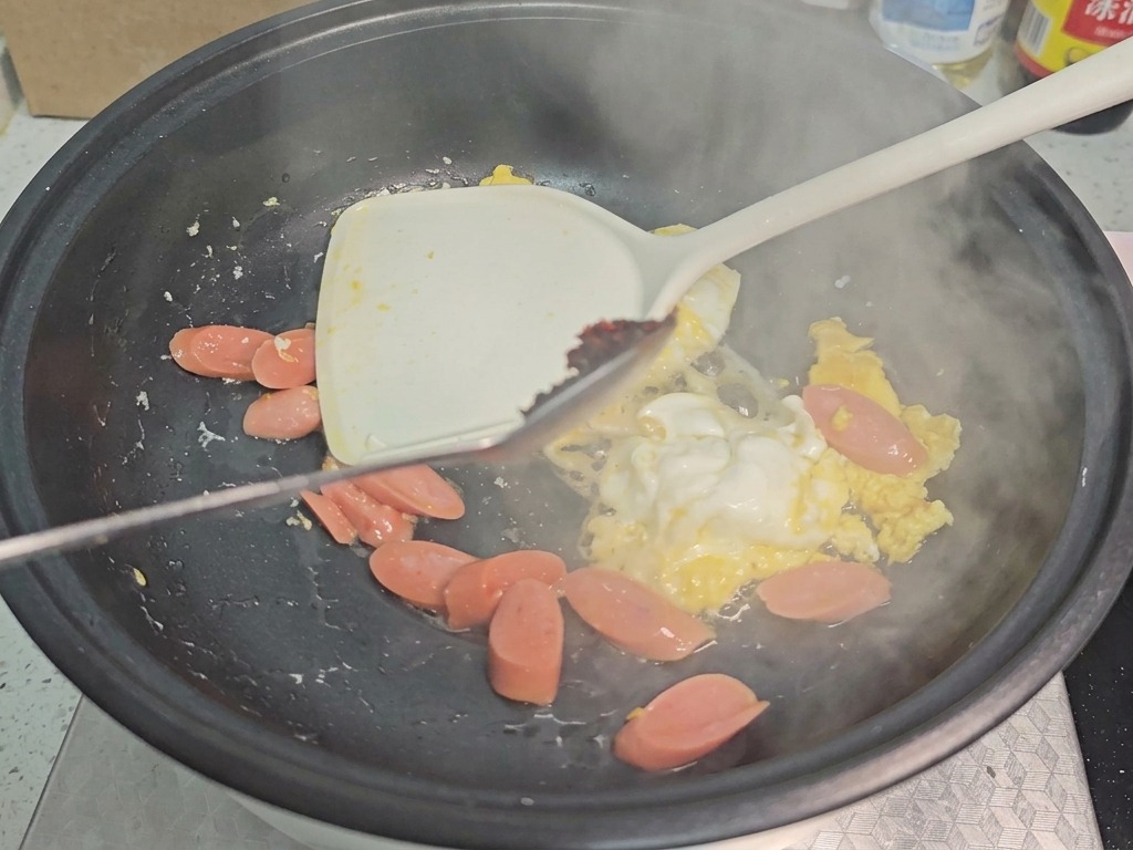 Ein Löffel dunkelrote Chilisauce wird unter das Rührei und die Würstchenscheiben in einer schwarzen Pfanne gemischt.