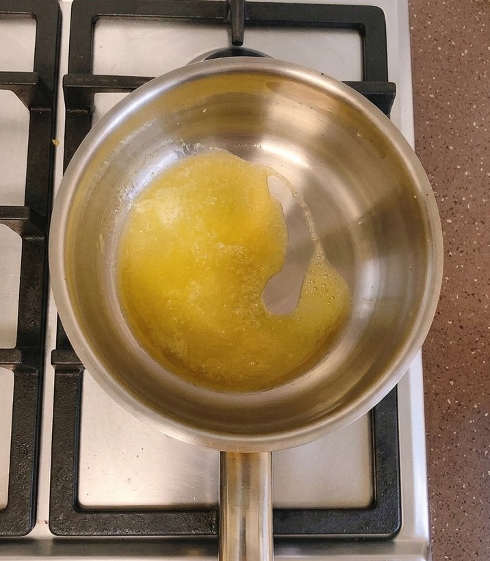 Ein Edelstahltopf auf einem Gasherd mit geschmolzener gelber Butter, in deren Mitte ein Löffel weißes Mehl gegeben wird.