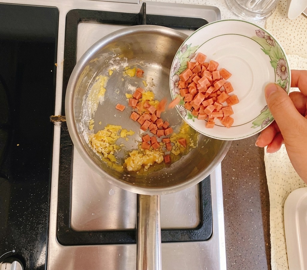 Gewürfelter Schinken wird von einem kleinen weißen Teller in einen Topf mit einer goldgelben Butter-Mehl-Mischung gegeben.