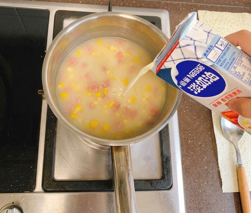 Flüssige Sahne wird aus einem blau-weißen Karton in einen köchelnden Topf mit cremiger Maissuppe auf dem Herd gegossen.