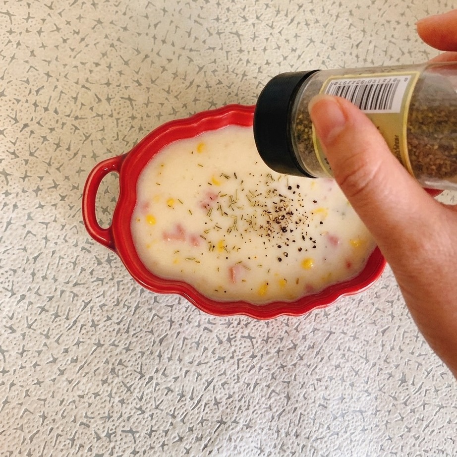 Eine Hand hält ein Gewürzglas und streut getrocknete Kräuter über eine rote Schüssel mit cremiger Schinken-Mais-Suppe.