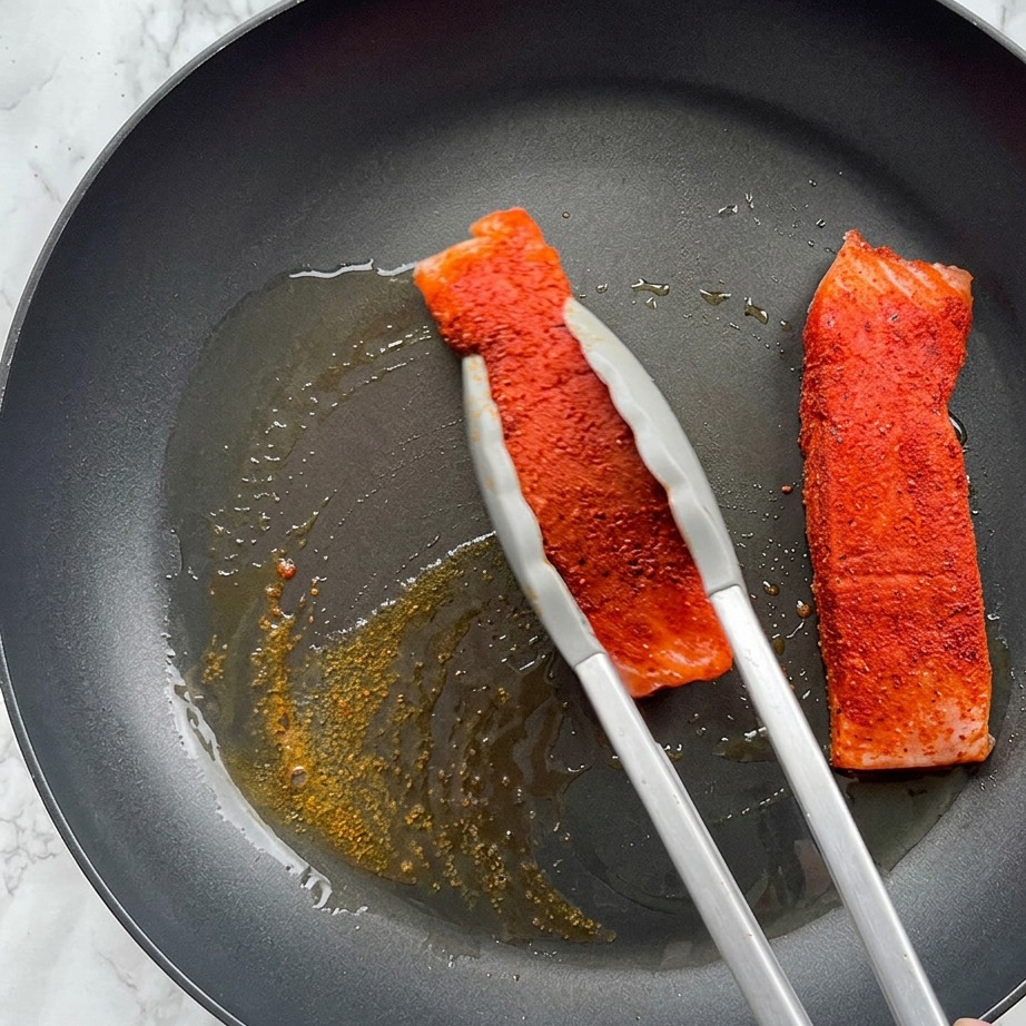 Ein gewürztes Lachsfilet wird in einer heißen Pfanne mit schäumendem Olivenöl angebraten und mit einer Metallzange gehalten.