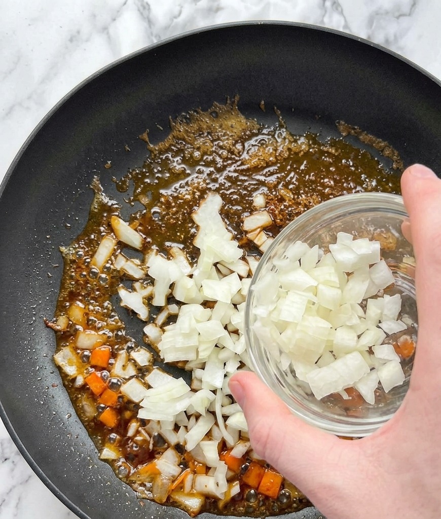 Gewürfelte weiße Zwiebeln werden aus einer Glasschüssel in eine Pfanne mit dem Bratensatz des Lachses gegeben.