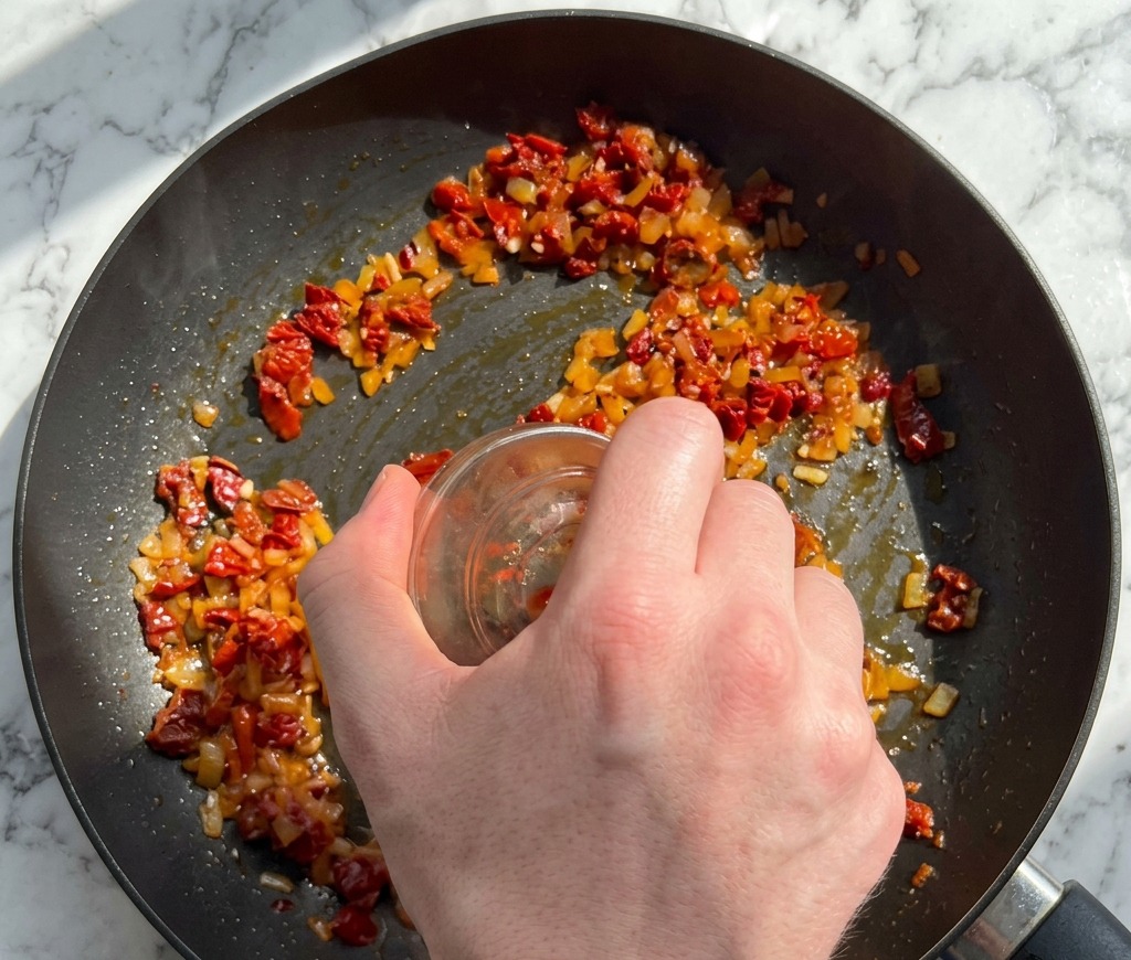 Eine Hand gibt gehackte getrocknete Tomaten und Chiliflocken aus einer kleinen Glasschüssel zu den angeschwitzten Zutaten in die Pfanne.