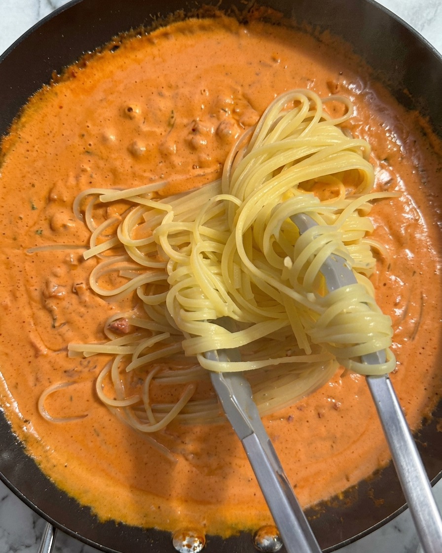 Eine Portion gekochte Nudeln wird mit einer Zange angehoben und in die Pfanne mit der cremigen Orangensauce gegeben.