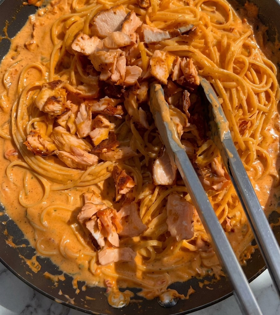 Stücke von gegartem rosa Lachs werden mit einer Metallzange zur cremigen Pasta in die Pfanne gegeben.