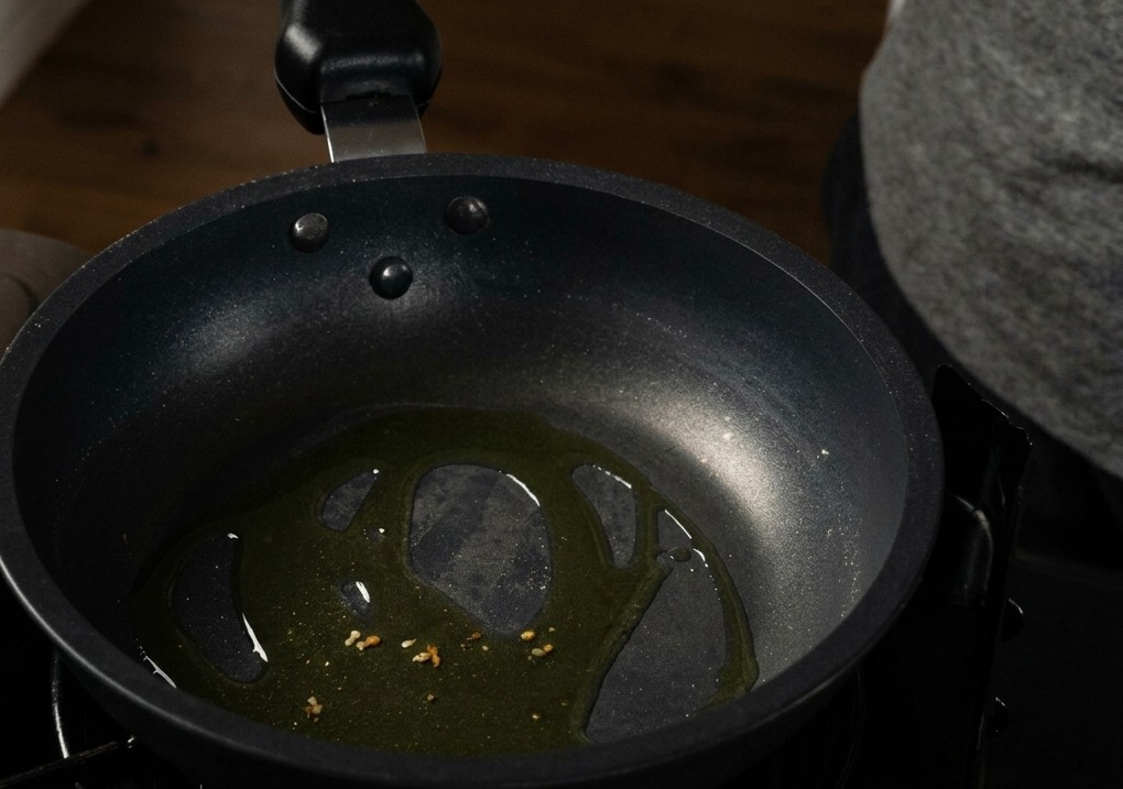 Gehackte Knoblauchstücke brodeln in Olivenöl am Boden einer schwarzen, beschichteten Bratpfanne.
