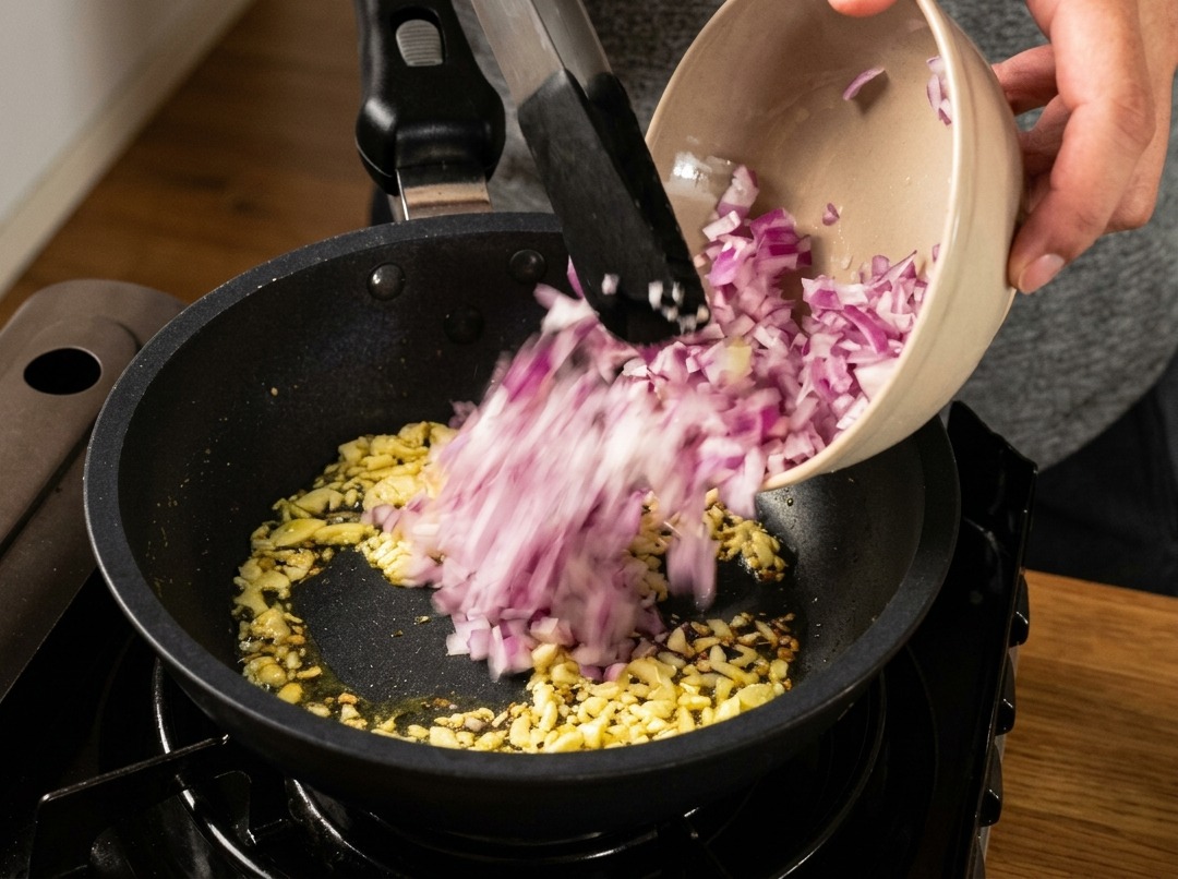 Gewürfelte rote Zwiebeln werden aus einer Schüssel in eine Bratpfanne gegeben, um sie mit Knoblauch anzuschwitzen.