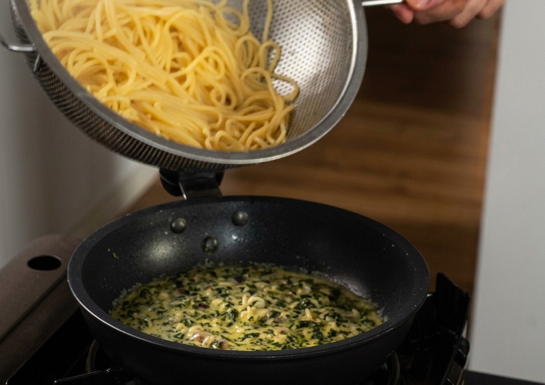 Gekochte Spaghetti werden aus einem Edelstahlsieb in eine Pfanne mit cremiger, mit grünem Basilikum durchzogener Sauce gegeben.