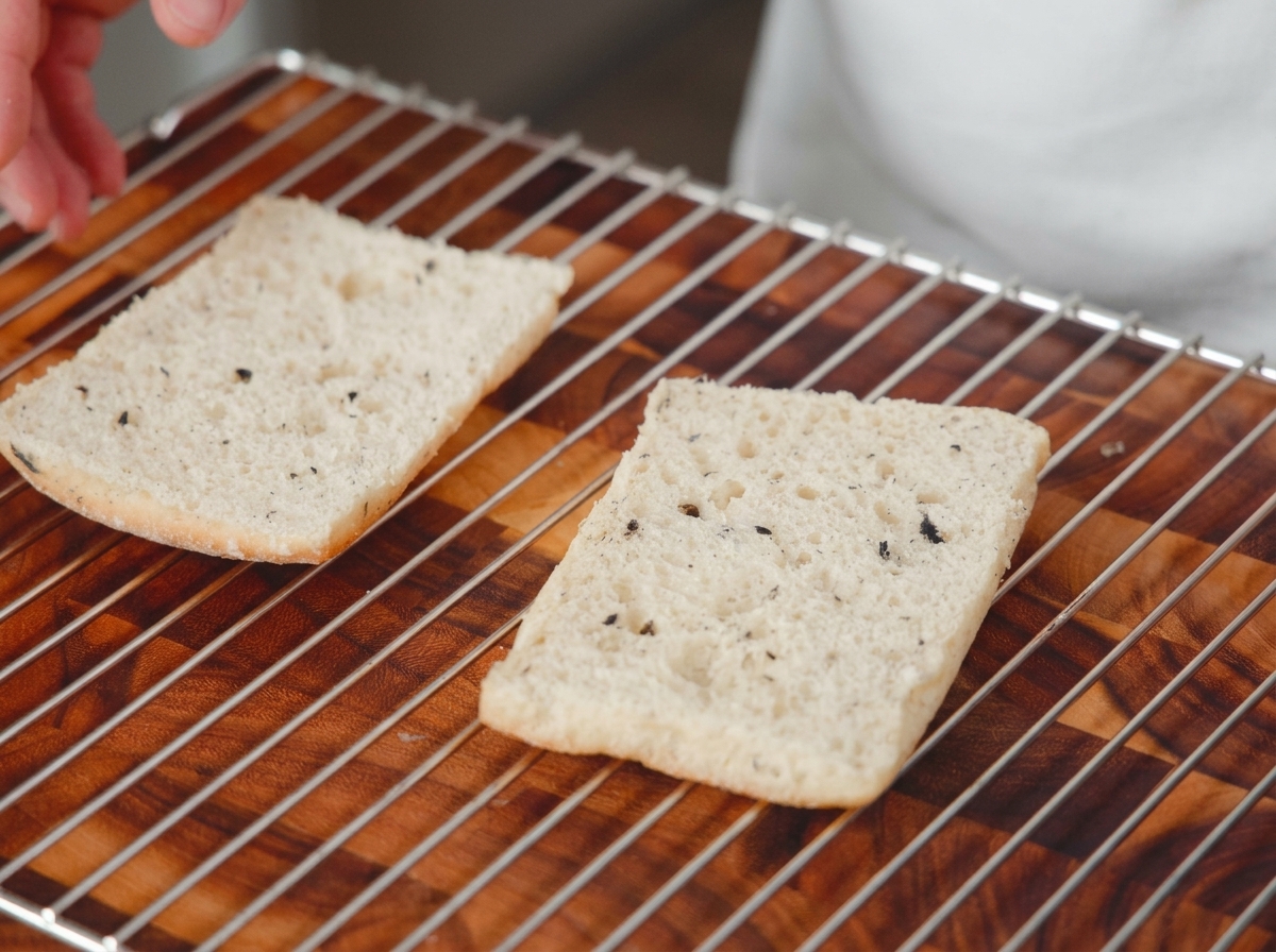 Zwei Hälften geschnittenes Brot liegen auf einem Ofenrost über einer Holzoberfläche und sind bereit zum Rösten.