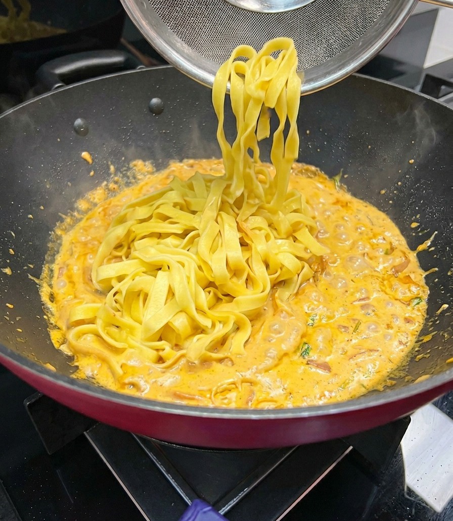 Breite Fettuccine-Nudeln werden mit einem Sieblöffel angehoben und in eine Pfanne mit köchelnder, orangefarbener Cremesauce gegeben.