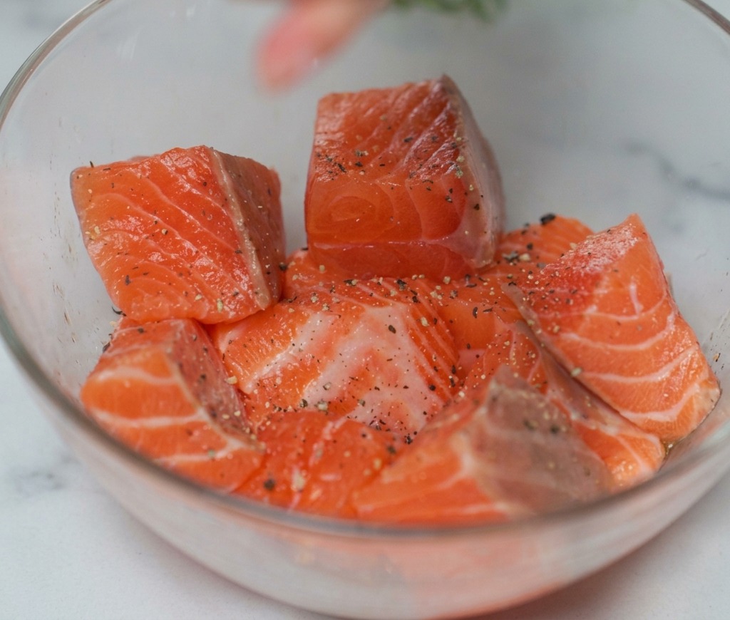 Rohe Lachswürfel in einer Glasschüssel werden mit Salz und schwarzem Pfeffer aus einer Mühle bestreut.