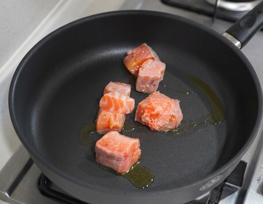 Lachswürfel braten in einer schwarzen, beschichteten Pfanne mit Olivenöl auf einem Gasherd.