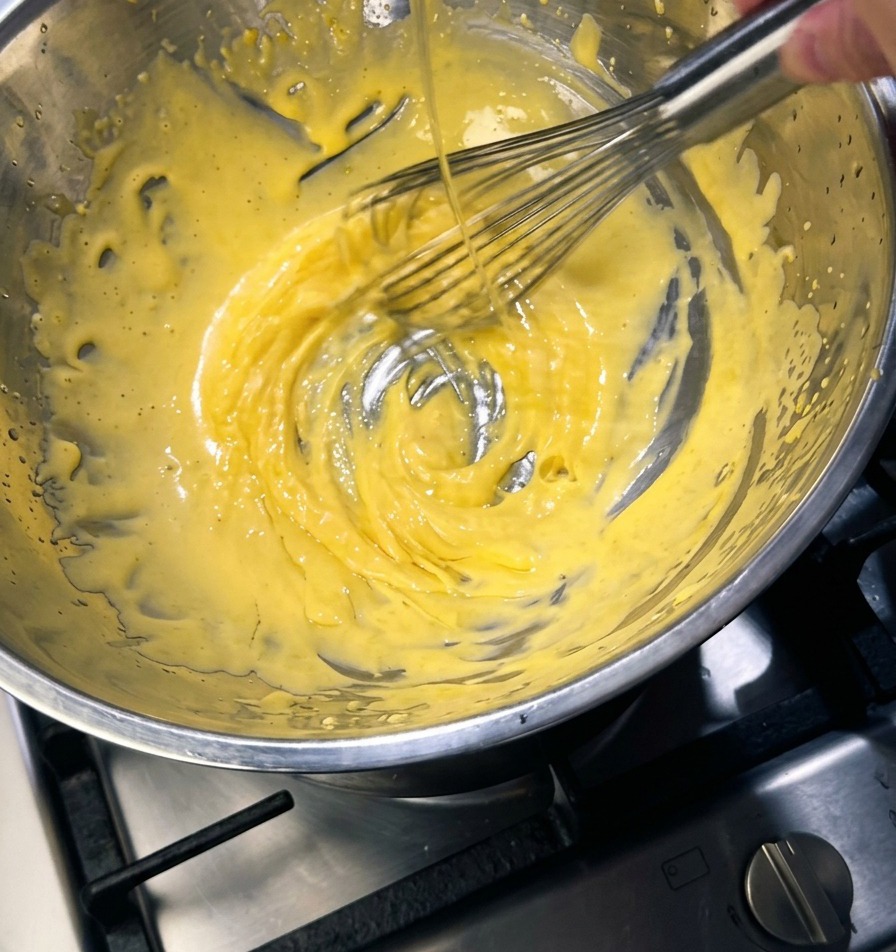 Ein Schneebesen rührt eine dicke, hellgelbe Masse in einer Metallschüssel, während ein dünner Strahl goldener Butter hineingegossen wird.