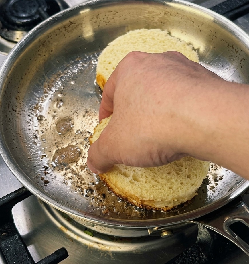 Eine Hand drückt zwei dicke, runde Scheiben Brioche-Brot in einer Pfanne flach an, um eine goldbraune Kruste zu erzeugen.
